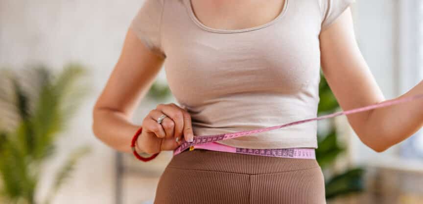 Frau misst ihren Taillenumfang mit einem rosa Maßband