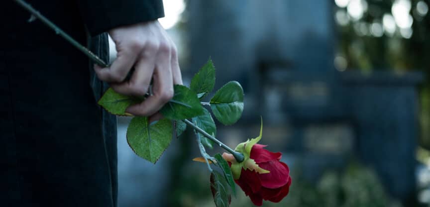 Hand hält eine rote Rose auf einem Friedhof als Zeichen der Trauer