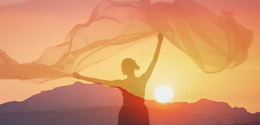 Silhouette einer Frau mit wehendem Tuch vor einem Sonnenuntergang in den Bergen.