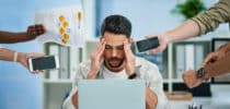 Gestresster Mann im Büro sitzt vor seinem Laptop und hält sich den Kopf, während mehrere Hände ihm Dokumente, Smartphones und Anweisungen entgegenstrecken.