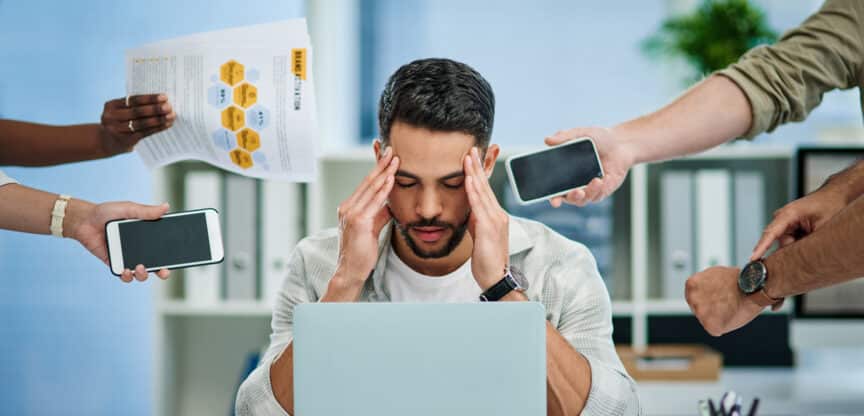 Gestresster Mann im Büro sitzt vor seinem Laptop und hält sich den Kopf, während mehrere Hände ihm Dokumente, Smartphones und Anweisungen entgegenstrecken.