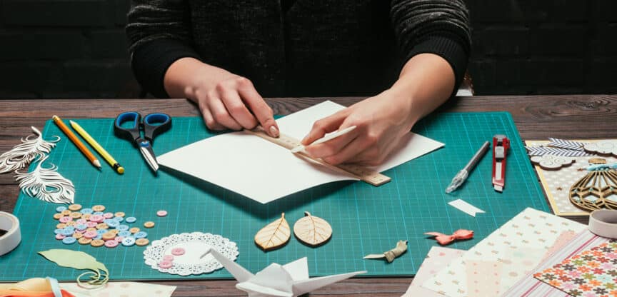 Person arbeitet an einem Bastelprojekt mit Schere, Lineal, Papier und Dekorationen auf einem Schneidematten-Untergrund.