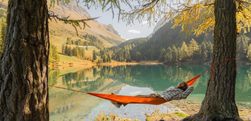 Person entspannt in einer orangefarbenen Hängematte zwischen zwei Bäumen am Ufer eines kristallklaren Bergsees, umgeben von herbstlicher Natur.