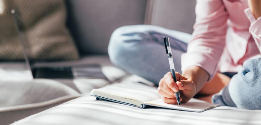 Person sitzt auf einer Couch und schreibt mit einem Stift in ein Notizbuch, während ein Laptop im Hintergrund liegt.