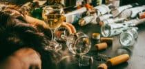 Person mit gesenktem Kopf neben leeren Alkoholflaschen und Weingläsern auf einem Tisch.
