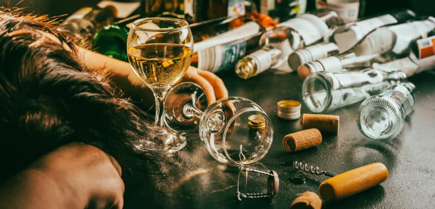 Person mit gesenktem Kopf neben leeren Alkoholflaschen und Weingläsern auf einem Tisch.