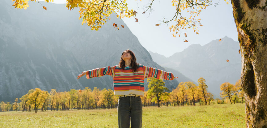 Frau mit buntem Pullover steht glücklich in einer Berglandschaft.