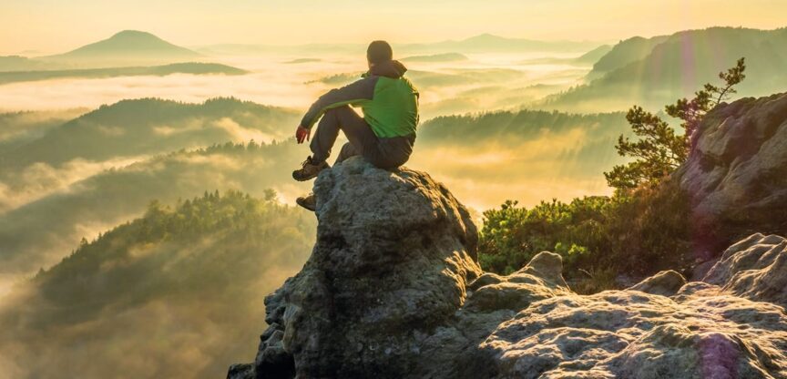 Bergwanderer sitzt auf Felsvorsprung in der Sonne und blickt auf im Nebel liegende Täler.
