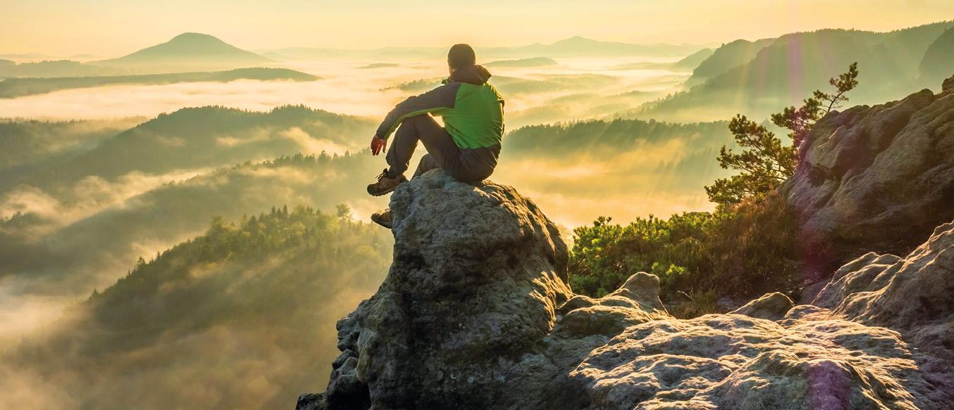Bergwanderer sitzt auf Felsvorsprung in der Sonne und blickt auf im Nebel liegende Täler.