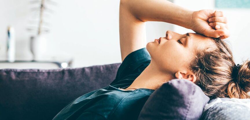 Junge Frau liegt mit geschlossenen Augen auf Sofa, Hand auf der Stirn.