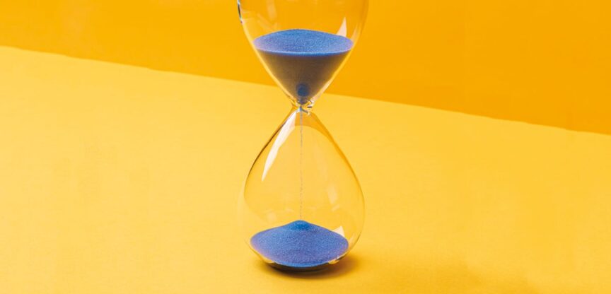 Sanduhr mit blauem Sand vor gelbem Hintergrund.