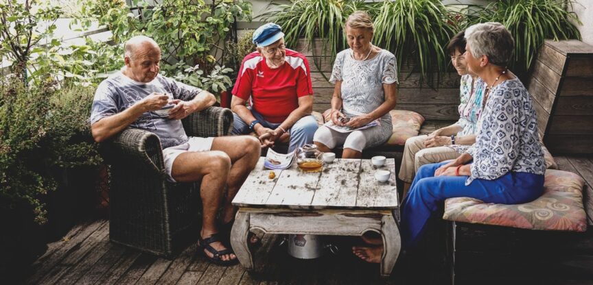 Fünf ältere Männer und Frauen sitzen um Couchtisch und reden.
