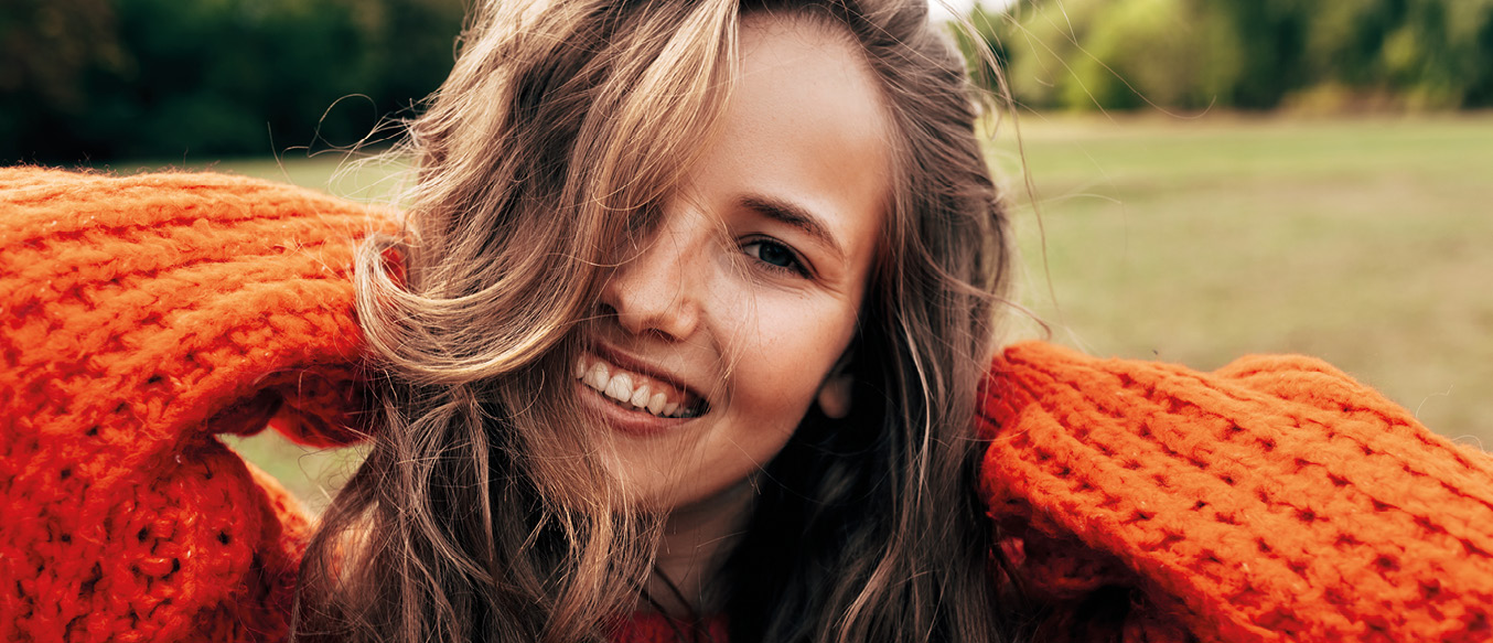 Junge Frau im orangen Strickpullover fasst lächelnd ihre langen brünetten Haare im Nacken zusammen.
