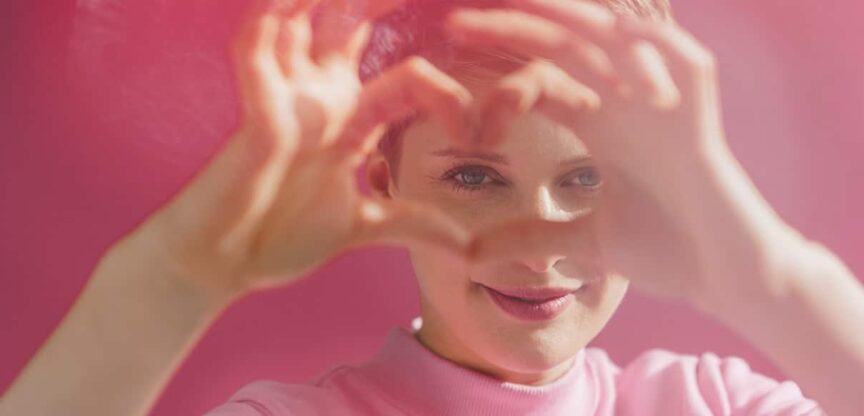 Lächelnde Frau vor rosa Hintergrund macht mit den Fingern ein Herz vor ihrem Gesicht.
