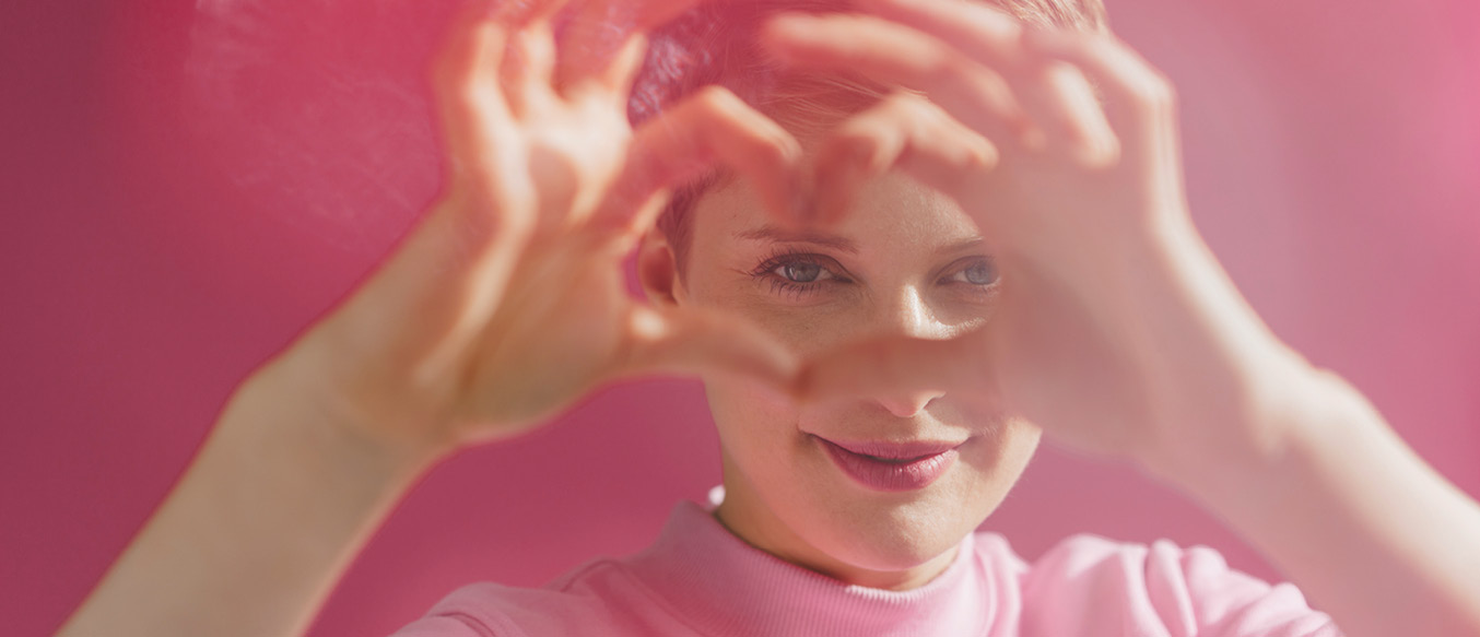 Lächelnde Frau vor rosa Hintergrund macht mit den Fingern ein Herz vor ihrem Gesicht.