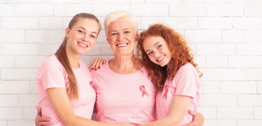 Zwei junge und eine ältere Frau mit rosa Pink-Ribbon-T-Shirts stehen lächelnd eng beisammen.