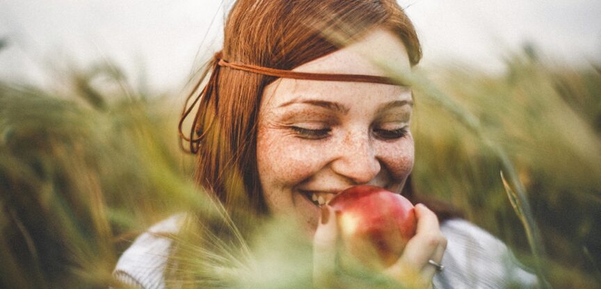 Lächelnde rothaarige junge Frau mit Sommersprossen mit einem Apfel in einem Kornfeld.