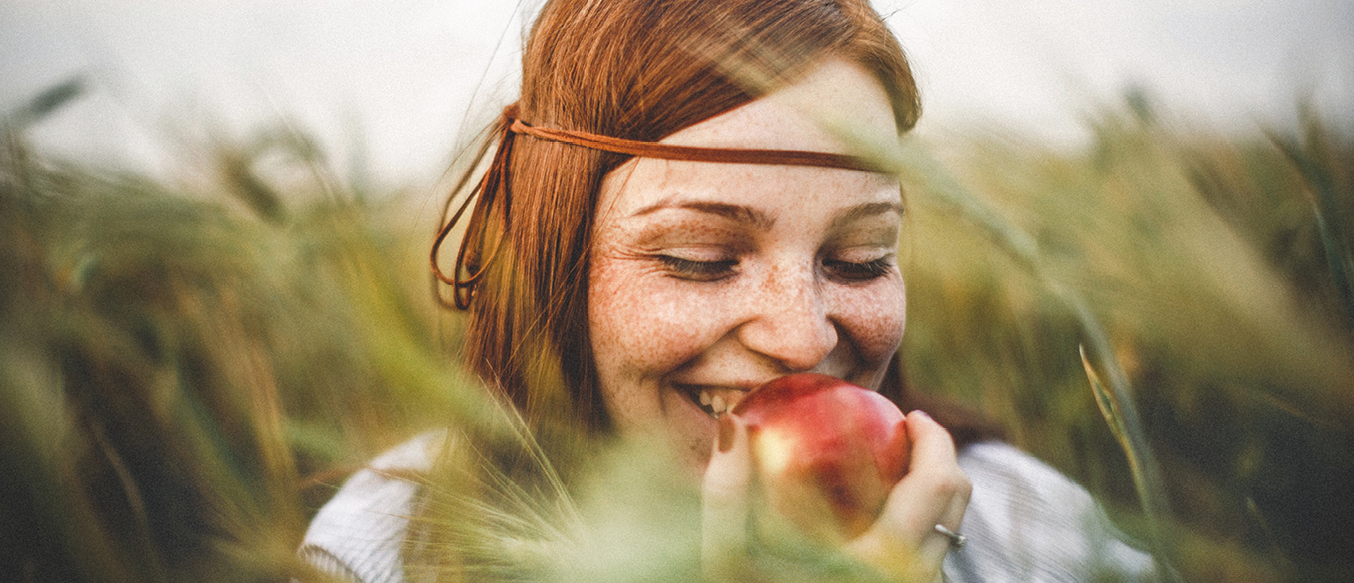 Lächelnde rothaarige junge Frau mit Sommersprossen mit einem Apfel in einem Kornfeld.