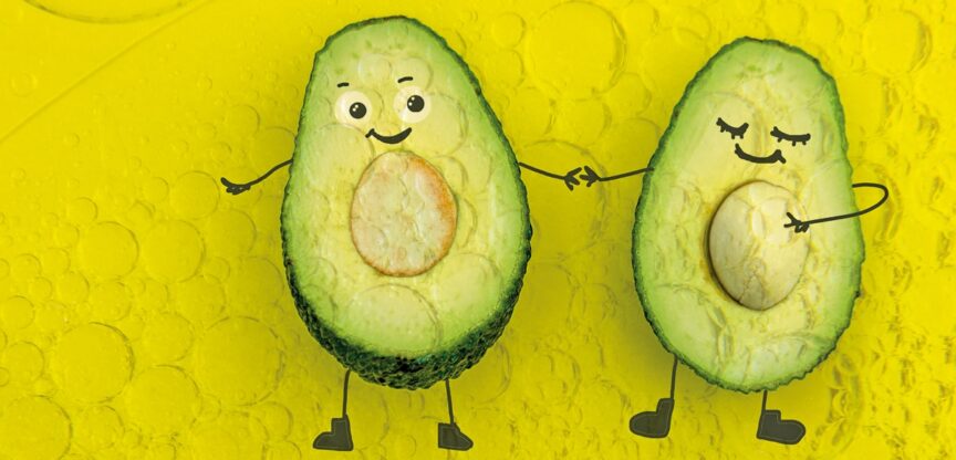 Zwei Avocado-Hälften auf gelbem Grund mit lustigen aufgemalten Gesichtern halten Händchen.