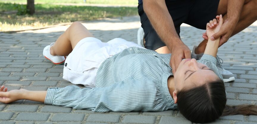 Junge Frau liegt ohnmächtig am Boden, Mann fühlt ihren Puls.