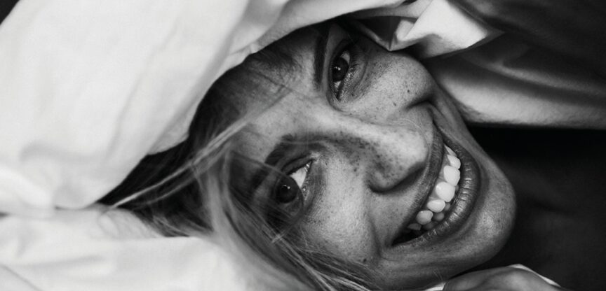 Lachende junge Frau im Bett hält sich Decke über den Kopf.