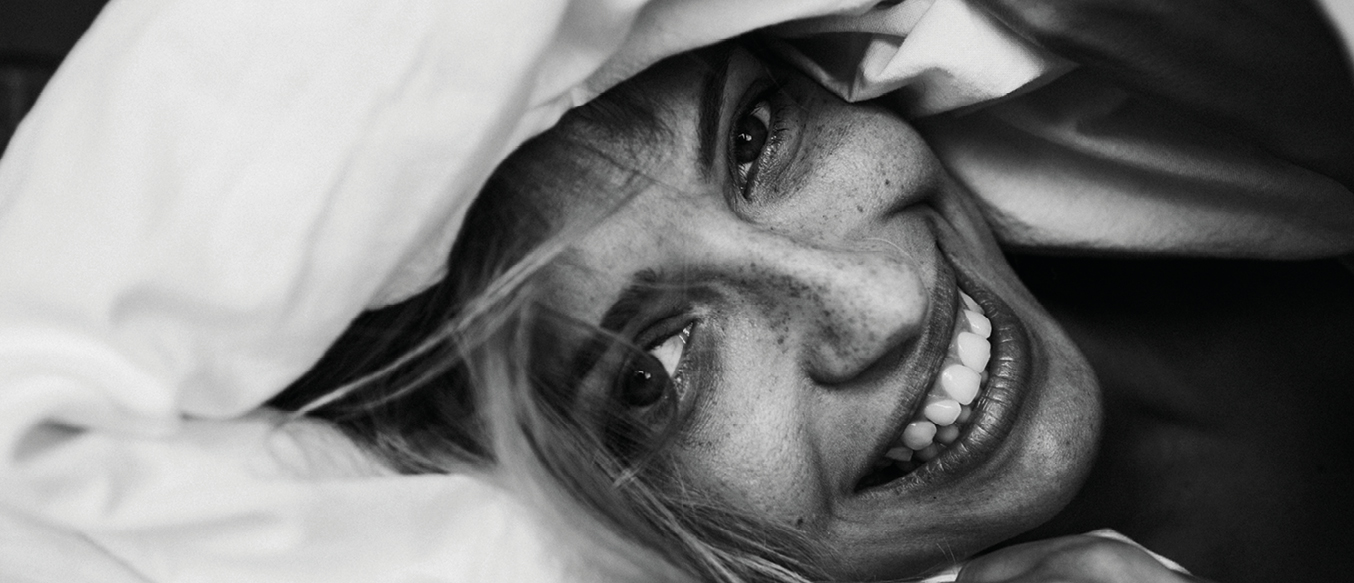 Lachende junge Frau im Bett hält sich Decke über den Kopf.