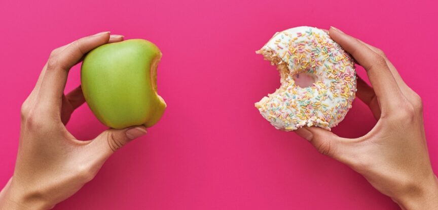 Zwei Frauenhände auf pinktem Grund halten angebissenen grünen Apfel und angebissenen Donut.