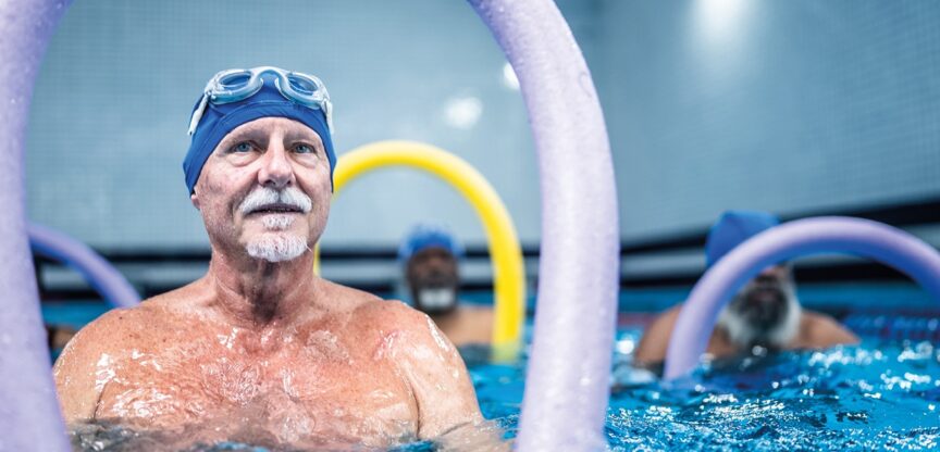 Älterer Mann mit Badehaube, Schwimmbrille und Wassernudel bis zur Brust im Schwimmbecken bei der Aquagymnastik.