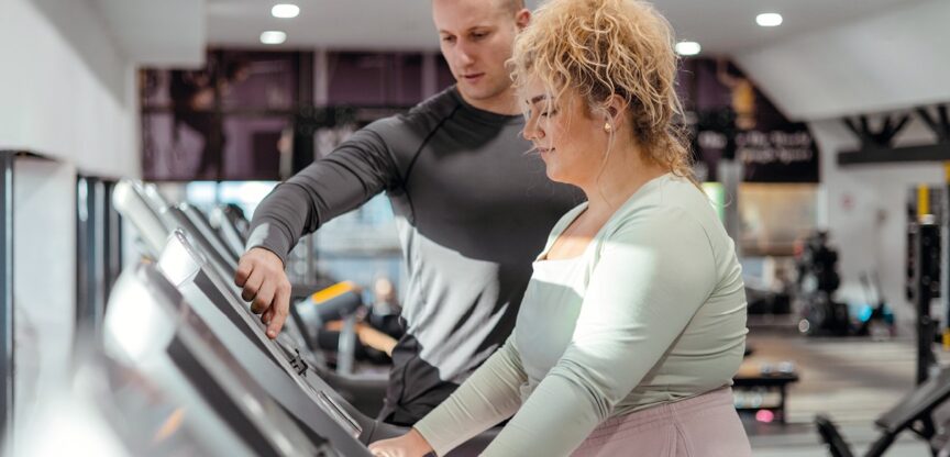 Übergewichtige Frau am Fitnessgerät mit Trainer.