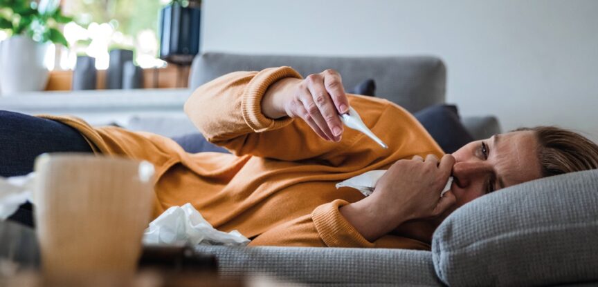 Frau mit oranger Jacke liegt auf grauem Sofa und blickt auf Fieberthermometer.