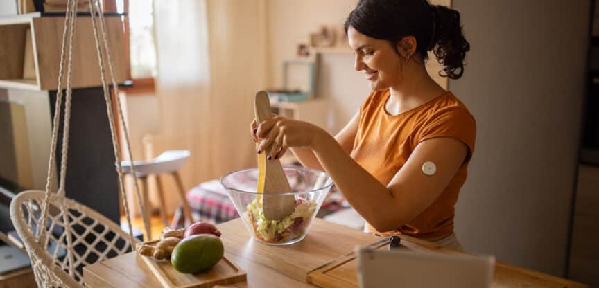 Eine junge Frau mit Glukosesensor am Arm bereitet in ihrer Küche einen Salat zu.