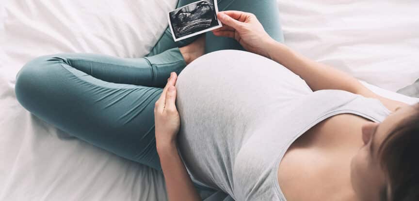 Schwangere Frau sitzt im Bett, hält eine Ultraschallaufnahme in der Hand und berührt sanft ihren Babybauch.