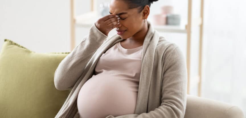 Schwangere Frau sitzt auf einem Sofa, hält sich den Kopf vor Schmerzen oder Erschöpfung und stützt ihre Hand auf den Bauch.