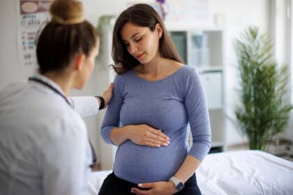 Ärztin spricht mit einer schwangeren Frau, die sich den Bauch hält und nachdenklich wirkt, in einer hellen Arztpraxis.