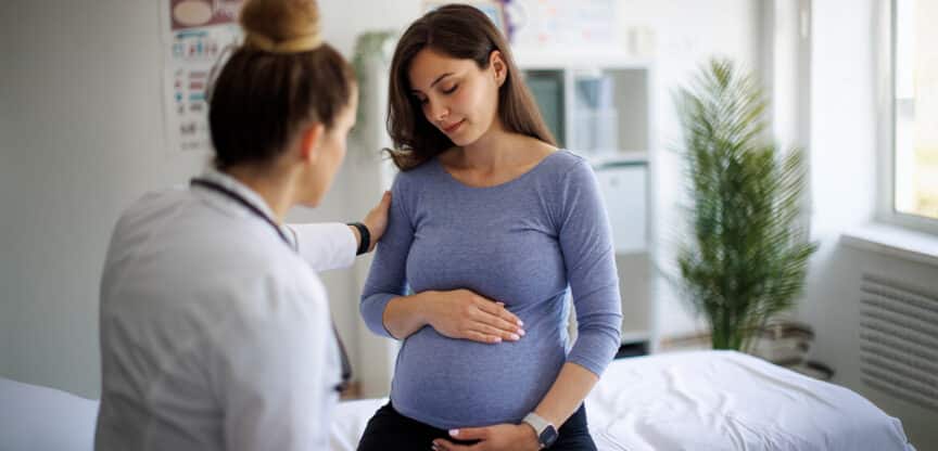 Ärztin spricht mit einer schwangeren Frau, die sich den Bauch hält und nachdenklich wirkt, in einer hellen Arztpraxis.