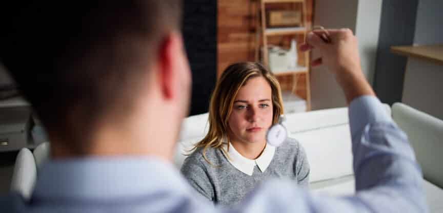 Therapeut führt Hypnose mit einer jungen Frau durch, die konzentriert auf ein Pendel blickt.