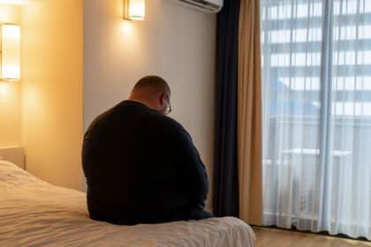 Übergewichtiger Mann sitzt nachdenklich am Bettrand in einem abgedunkelten Zimmer mit Blick nach draußen – Symbolbild für Einsamkeit oder mentale Belastung.