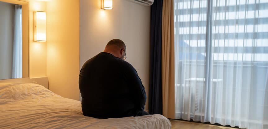 Übergewichtiger Mann sitzt nachdenklich am Bettrand in einem abgedunkelten Zimmer mit Blick nach draußen – Symbolbild für Einsamkeit oder mentale Belastung.