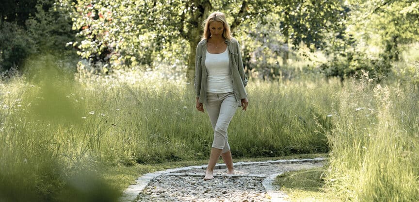 Frau geht barfuß auf einem Naturpfad aus Steinen durch eine grüne Wiese mit Bäumen – symbolisiert Achtsamkeit, Entspannung und Verbindung zur Natur.