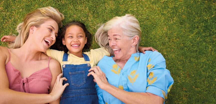 Drei Generationen einer Familie – Großmutter, Mutter und Tochter – liegen lachend auf einer Wiese und genießen gemeinsame Zeit.