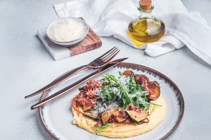 Cremige Polenta mit italienischem Gemüse, Rucola und geriebenem Parmesan, angerichtet auf einem Teller mit goldenem Besteck – daneben Olivenöl und eine Schale Käse.