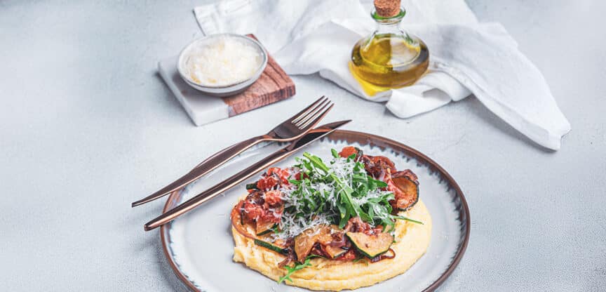 Cremige Polenta mit italienischem Gemüse, Rucola und geriebenem Parmesan, angerichtet auf einem Teller mit goldenem Besteck – daneben Olivenöl und eine Schale Käse.