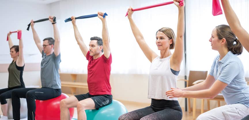 Gruppe Erwachsener beim physiotherapeutisch angeleiteten Training mit Gymnastikbändern auf Sitzbällen; Therapeutin unterstützt gezielte Bewegungsübungen für Mobilität und Kraft.