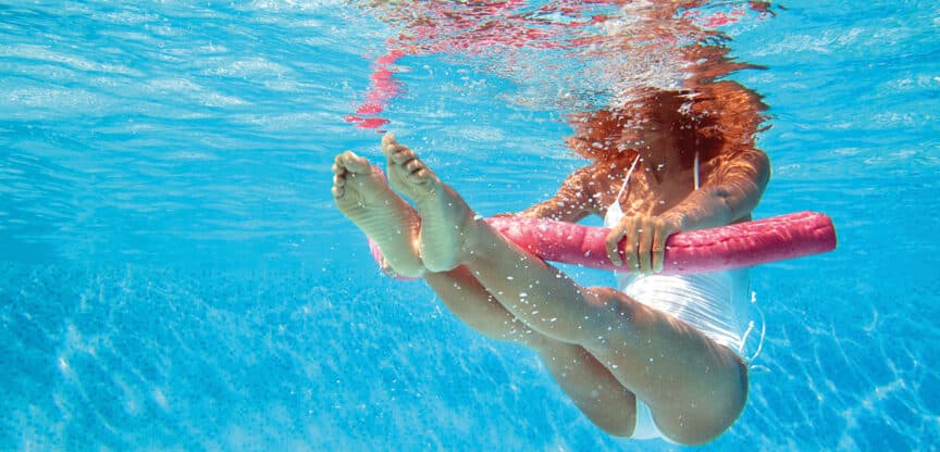 Unterwasseraufnahme einer Frau in weißem Badeanzug, die mit einer pinken Schwimmnudel Aqua-Fitnessübungen im Pool macht.