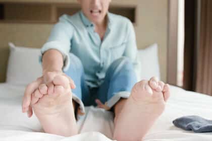 Mann sitzt auf einem Bett, greift sich mit schmerzverzerrtem Gesicht an die Füße – Symbol für Fußkrämpfe oder Beschwerden.