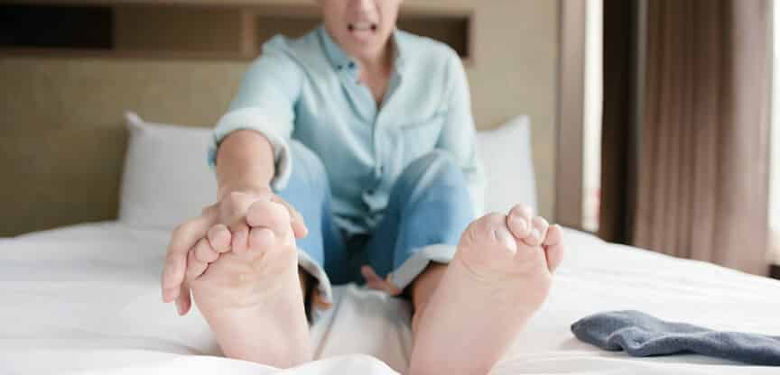 Mann sitzt auf einem Bett, greift sich mit schmerzverzerrtem Gesicht an die Füße – Symbol für Fußkrämpfe oder Beschwerden.