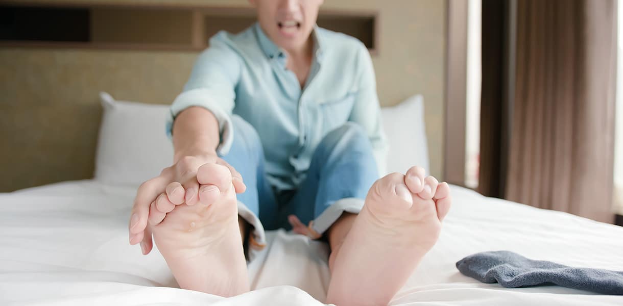 Mann sitzt auf einem Bett, greift sich mit schmerzverzerrtem Gesicht an die Füße – Symbol für Fußkrämpfe oder Beschwerden.