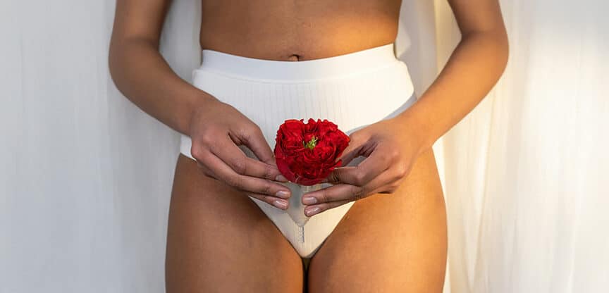 Frau in Unterwäsche hält eine rote Blume symbolisch vor ihrem Unterleib.