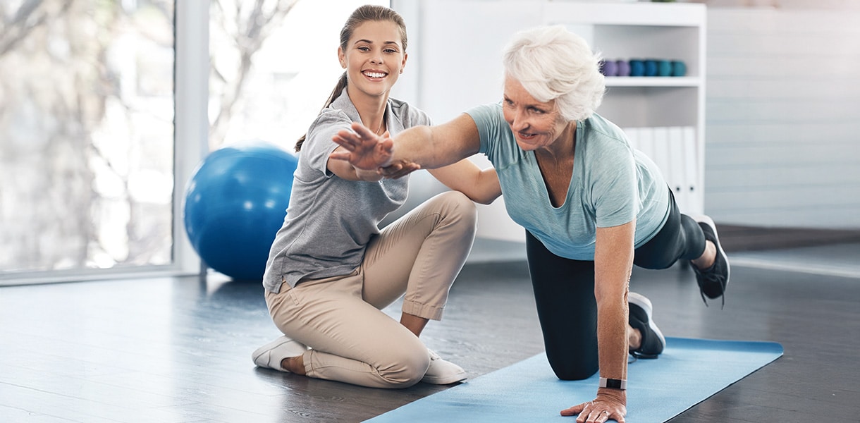 Ältere Frau macht Gleichgewichtsübung auf Matte, während eine Trainerin sie unterstützt und motiviert.