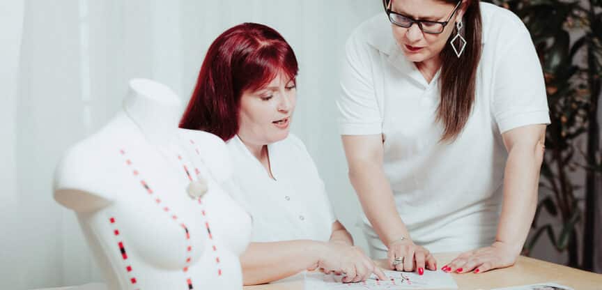 Zwei Frauen in weißer Kleidung besprechen gemeinsam ein Brustmodell und Unterlagen zur Brustkrebsfrüherkennung.